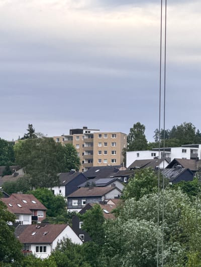 The image shows a view of residential buildings in Bad Berleburg, Germany. In the background, a tall, beige apartment building with a dark roof is visible, featuring multiple floors with evenly spaced windows. In front of this, a row of smaller, white and dark-roofed buildings are situated. Closer to the foreground, several houses with pitched roofs, some of which are covered with solar panels, are nestled among lush green trees. The sky is overcast and appears pale blue and white. Two thin, vertical metallic poles run through the right side of the frame, suggesting the presence of utility lines or scaffolding. There are no people visible in the image.