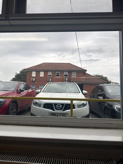 The scene is a static exterior view captured from an indoor window. The immediate foreground shows a white window frame, sill, and a radiator grill. A thin dark cable hangs vertically in the upper-left portion of the windowpane, which also exhibits horizontal light reflections.

In the midground, a parking area features three parked vehicles. From left to right: a red SUV, a white Nissan Qashqai with the UK license plate "PN12 FRC", and a black car, possibly a Mercedes-Benz. A yellow horizontal bar or barrier runs across the lower front sections of the vehicles.

The background consists of a two-story brick building with a red tiled roof, multiple white-framed windows, and at least two external air conditioning units mounted on the brick wall. Trees are visible around and behind the building. The sky is uniformly overcast. No individuals or active interactions are present. This scene is located in Boston, United Kingdom.