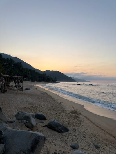 A serene beach scene unfolds at twilight. Gentle waves kiss the sandy shore, which is dotted with large, grey boulders. Lush green mountains rise in the background, silhouetted against a soft, pastel-colored sky with hues of orange, pink, and grey. A simple wooden structure stands near the edge of the vegetation. The overall mood is peaceful and calming.