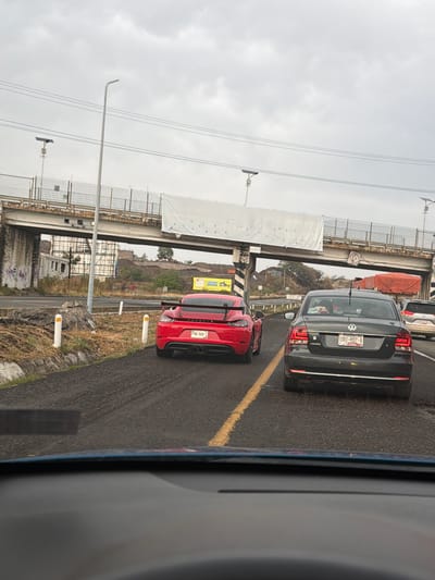 The image depicts a congested roadway in San Bernardino Chalchihuapan, Mexico, under an overcast sky. Visible are several vehicles, including a red sports car, a gray sedan, and at least one other vehicle partially visible ahead of the sedan. The vehicles are traveling on a paved road with a yellow lane divider. A bridge structure with metal framework and concrete supports runs overhead. A weathered billboard can be seen in the background near a hillside. White bollards line the side of the road nearest the sports car. The view appears to be taken from inside a vehicle, looking forward through the windshield.