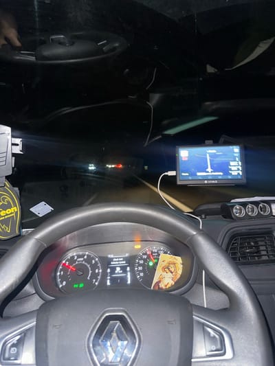 The image depicts the interior of a Renault vehicle at night, viewed from the driver's perspective, located in Oostkamp, Belgium. The vehicle is in motion, with the speedometer indicating approximately 65 km/h and the tachometer at approximately 2000 RPM. The dashboard's central display shows the time as 18:19 and displays a "Check Anti-Pollution System" warning, accompanied by an active orange warning light. Green indicator lights for low beam headlights are also illuminated. A laminated religious icon, likely depicting Mary and Jesus, is tucked into the speedometer dial. To the left, a yellow "Areon" brand air freshener is partially visible.

A VIVAS-branded electronic navigation device, displaying a map and route, is mounted on the dashboard to the right and is connected via a white USB cable. Adjacent to the navigation device on the right, a black accessory unit with three illuminated circular ports (likely 12V power outlets) is present. A human hand is partially visible in the upper left section of the image, above the dashboard. Through the windshield, a dark road is visible, illuminated by vehicle headlights, with distant red taillights and white headlights of other vehicles indicating traffic. The interior is primarily dark, illuminated by dashboard lights and screens.