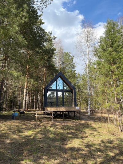 The image depicts a modern A-frame cabin situated within a forest clearing in Мишкино, Russia. The cabin, constructed with a dark grey frame and light wooden siding, features a distinctive triangular shape with large, expansive glass windows on its front facade, reflecting the blue sky and surrounding pine trees. It stands on an elevated wooden deck, beneath which additional wooden supports are visible. A black metal railing lines the deck, where a dark metal outdoor table and chair are visible. A white plaque bearing the number "20" is affixed to the cabin's exterior near the deck.
The surrounding environment is a dense forest, dominated by tall coniferous trees, primarily pines with their characteristic green needles. Scattered among them are several deciduous trees, notably birches, identifiable by their white bark and bare branches, suggesting late winter or early spring. The ground is covered with dry grass and patches of earth, with numerous tree shadows cast across it from the bright sunlight. Near the base of the cabin, a blue plastic bag, possibly a garbage bag, rests on a black stand.
The scene appears to be captured during the daytime, under a bright sky with scattered white cumulus clouds. There are no people visible, and no specific activity is taking place, presenting a tranquil and still moment of a modern dwelling integrated into a natural, forested landscape.
