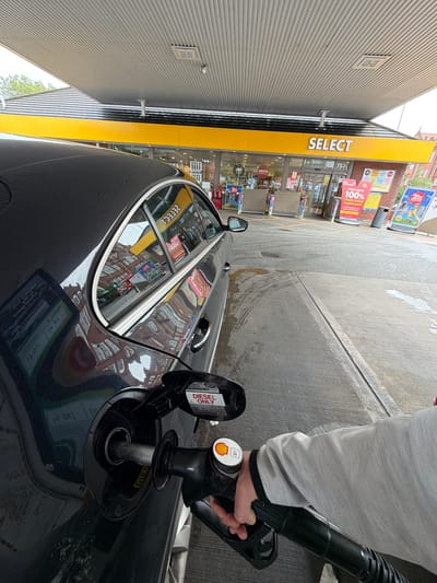 A photograph, rotated 90 degrees clockwise, captures a person refueling a dark-colored car with diesel at a Shell petrol station in Leicester, United Kingdom. The black fuel nozzle, marked with a yellow Shell logo and "B7," is inserted into the car's fuel tank, which has an open flap bearing a "DIESEL ONLY" sticker. The arm and hand of the person, clad in a light grey long-sleeved top, are visible holding the nozzle.
Adjacent to the car is a Shell Select convenience store, characterized by its bright yellow "SELECT" signage and large glass windows. Within the store's display, signs for "COSTA" and "deli" are visible. Outside, promotional posters advertise "Now up to 100% cleans critical engine parts" and "2 for..." offers.
The concrete forecourt shows areas of wetness and tire marks. The car's glossy surface and windows reflect the Shell Select store, its signage (including reversed "ГЕГЕСІ" for SELECT and "DIES" for DIESEL), and surrounding buildings. The scene appears to be set during daytime, under what seems to be an overcast sky.