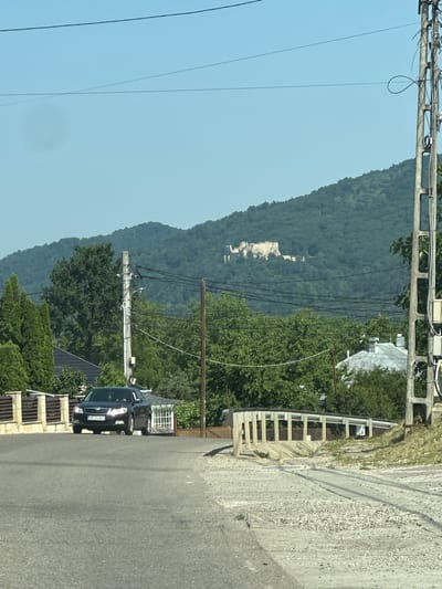 The image shows a view of a rural road in Targu Neamt, Romania. A black Skoda Octavia is driving on the road. In the background, a medieval fortress is visible on a forested hill. The sky is clear and blue. There are trees and houses on the sides of the road. A power pole with wires is on the right side of the image.
