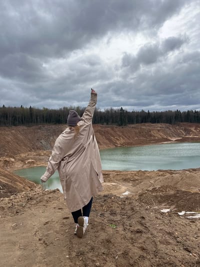The image shows a person standing on a dirt embankment overlooking a body of water. The person is wearing a beige trench coat, dark pants, a grey hat, and white sneakers. Their arms are outstretched towards the cloudy sky. On the left side of the image, a line of trees runs along the edge of the embankment. The location is near Ivlevo, Russia.
