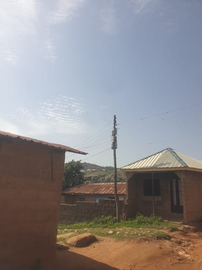 The image depicts an outdoor scene with a clear sky and scattered clouds. In the foreground, a structure made of reddish-brown material, possibly adobe or mud brick, with a corrugated metal roof is visible on the left. To the right, another building with a similar construction and a light-colored corrugated metal roof with a green trim is present. A wooden pole with electrical equipment and wires runs vertically in the middle ground, connecting to the buildings. Beyond these structures, there are more buildings with corrugated metal roofs and walls constructed from cinder blocks. In the background, a hilly landscape with rocks and vegetation can be seen. The ground in the foreground is a mix of dirt and scattered stones. The scene appears to be in a rural or semi-rural area. The image does not contain any elements that suggest it is located in the city of Fresno, United States of America.
