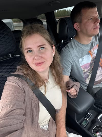 The media file shows two people, a man and a woman, sitting in the front seats of a car. The man is on the left, wearing a grey t-shirt with a graphic print and a seatbelt. He is looking upwards and appears to be relaxed. The woman is on the right, closer to the camera, wearing a light-colored shirt and a beige cardigan. She is also wearing a seatbelt and looking towards the camera with a slight smile. Her blonde hair is visible around her face.
The interior of the car is visible, including the dashboard with car keys, the center console, and the seats. The seats appear to be black leather with a quilted pattern. A seatbelt is clearly visible across the woman's chest. The background outside the car is blurred, but it appears to be a natural setting with trees.
The image is taken from a high angle, looking down on the subjects. The lighting suggests it might be daytime, possibly overcast, given the softness of the light. The overall mood seems relaxed and casual, possibly during a road trip. There is no visible text in the image.