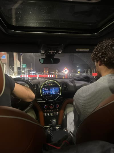 A view from inside a car, likely a Mini Cooper, facing forward through the windshield on a rainy night. A driver grips the steering wheel. The dashboard is visible with a lit-up navigation screen. Outside, a "40" speed limit sign is illuminated by streetlights. Toll booths with red and white striped poles are also visible through the rain-streaked windshield. The car's interior features brown leather seats. The reflection from the window is apparent.