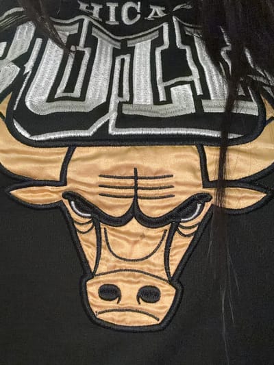 The image shows a close-up of an embroidered Chicago Bulls logo, featuring the team's iconic bull head and lettering, on a black garment. The bull's head is rendered in gold with black outlining, and its horns are depicted in gold. Above the bull's head, the word "CHICAGO" is embroidered in silver and white block letters. Black hair, likely from a person, partially obscures the top portion of the logo. The image composition is tight, focusing on the details of the embroidery and the texture of the fabric. There is no discernible environment or city context provided within the image itself.