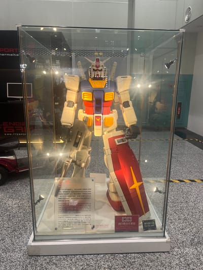 A large, multi-colored RX-78-2 Gundam model, standing upright and holding a beam rifle in its right hand with a shield on its left arm, is enclosed within a rectangular glass display case in Taoyuan City, Taiwan. The model features white, blue, red, and yellow components, consistent with the classic Gundam design. Multiple spotlights inside the case illuminate the model. A white informational plaque with Chinese and English text, likely detailing the Gundam, is positioned at the base of the model. Adjacent to it, a red sign displays "請勿觸碰或攀爬" (Please do not touch) and "非賣品" (Not for sale). The display case rests on a white base on a speckled grey floor. In the background to the left, a dark banner with partially visible text including "SPORT" and "TTESPORTS.COM.T" can be seen, along with a black monitor screen and the rear portion of a red mobility scooter. To the right, a light blue-green wall features a red fire extinguisher cabinet and other wall-mounted utilities. Yellow and black striped warning tape is visible on the floor to the right of the display case. Overhead lighting is reflected in the glass. No individuals are present in the immediate vicinity.