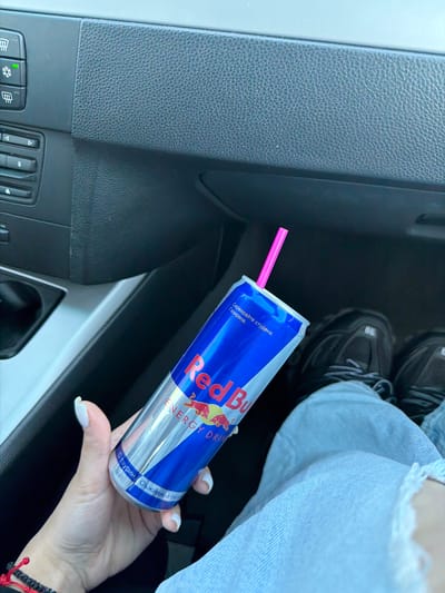 A right-handed person with pale skin and white manicured nails is holding a can of Red Bull with a pink straw in the interior of a car. The can of Red Bull is blue and silver with the words "Red Bull ENERGY DRINK" printed on it. The text on the can is in Bulgarian, including the phrases "Сервирайте студена. Газирана" which translates to "Serve cold. Carbonated" and "С таурин. Съживява тяло" which translates to "With taurine. Revitalizes the body." The hand is adorned with a black and red bracelet. Above the Red Bull, the person is wearing faded blue jeans and black and grey sneakers. The car interior is dark with grey and black textured panels. The location is assumed to be in Letnitsa, Bulgaria based on the Bulgarian text on the can.
