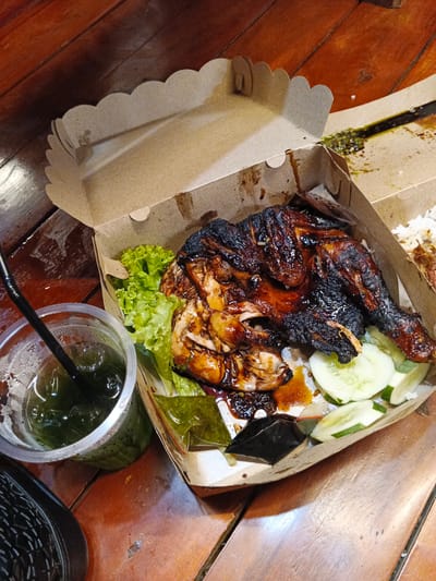 The image shows a takeout box containing a grilled chicken dish with rice, lettuce, and cucumber slices. A plastic cup with a green beverage and a black straw is placed next to the box. The meal is presented on a wooden table. The setting is likely a casual dining establishment in Kampongbaro, Indonesia, possibly a street food stall or a simple restaurant given the disposable packaging and wooden table. The lighting suggests an indoor setting, possibly during the evening or in a dimly lit area. There are no people visible in the image, and the focus is solely on the food and drink. The overall impression is one of a ready-to-eat meal.