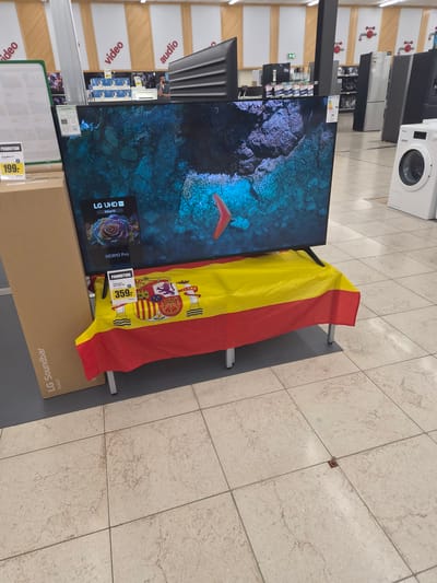 This media file captures a retail environment, likely an electronics store in Käerjeng, Luxembourg. The primary focus is on a large television display, showcasing a vibrant underwater scene. A Spanish flag is draped over the display stand, suggesting a promotional event or a national display.
In the foreground, a cardboard box for an "LG Soundbar" is visible, along with promotional pricing tags indicating "199-" and "359-". The television itself is identified as an "LG UHD AI 55UA73" supporting "HDR10 Pro". In the background, shelves stocked with various electronic items and appliances, including washing machines and refrigerators, are partially visible. The store appears well-lit, with overhead lighting illuminating the tiled floor. The overall scene conveys a commercial setting with a specific product promotion. There are no people clearly visible in the image.