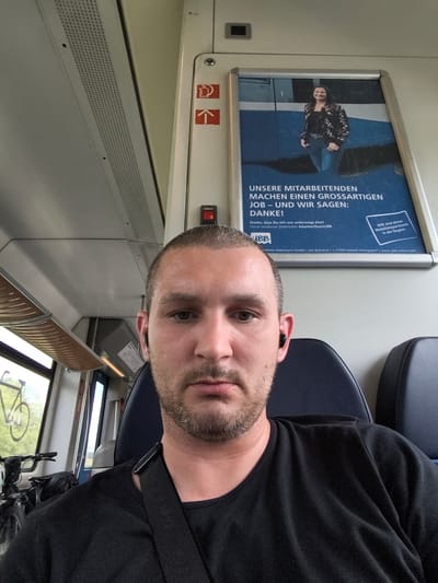 This media file shows a selfie taken by a man on a train. He is the primary subject, positioned in the foreground, looking directly at the camera with a neutral expression. He has short, buzzed hair and a light beard, and is wearing a black t-shirt and black earbuds. The background reveals the interior of a train carriage. To the left, there are windows showing greenery and a bicycle rack with a bicycle, suggesting a journey through a rural or suburban area. Above the man's head, the train's ceiling is visible, with perforated panels and lighting.

To the right of the man, a large advertisement is displayed on the train wall. The advertisement features a woman smiling and standing, with text in German. The visible text from the advertisement includes "UNSERE MITARBEITENDEN MACHEN EINEN GROSSARTIGEN JOB - UND WIR SAGEN: DANKE!" which translates to "OUR EMPLOYEES MAKE A GREAT JOB - AND WE SAY: THANK YOU!". It also mentions "Deine Usedomer Bäderbahn #starkesTeamUBB" and "WIR sind deine Mobilitätspartnerin in der Region", indicating that the advertisement is for the Usedomer Bäderbahn, a regional transport company. The location context of Züssow, Germany, places this scene within that region. The lighting suggests it is daytime. The overall atmosphere is casual, likely a commuter or leisure journey.
