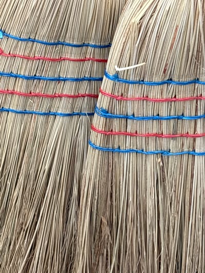 This is a close-up, detailed, forensic photograph of the bristles and binding of two brooms. The brooms are made of natural, straw-like material and are bound together with red and blue strings. The strings are wrapped horizontally around the broom heads, creating distinct bands. The bristles are thin and numerous, with a predominantly light brown and beige color. Some stray pieces of dried plant matter are visible among the bristles. The image is taken from a slightly angled perspective, focusing on the texture and construction of the brooms. The setting is not discernible beyond the objects themselves.