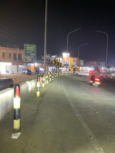 The image depicts a nighttime street scene in an urban or semi-urban environment, likely in Seuneubok Teungoh, Indonesia. The asphalt road, which appears to curve slightly to the right, is illuminated by various light sources.
Along the right side of the road, a series of black and yellow (or red) striped bollards or traffic barriers separate the thoroughfare from a paved curb. Some white debris is visible on the ground near these barriers. On the road itself, a blurred motorcycle with distinct red taillights is captured in motion, indicating ongoing traffic.
The left side of the street features multiple buildings, likely housing shops or businesses, marked by their illuminated windows and signs. Overhead, several streetlights with long, curved arms cast light onto the scene against the dark night sky. Utility poles and overhead power lines are also present. Among the visible signage is a prominent green billboard displaying the text "KUB Syariah Bank Aceh" along with the slogan "Hidup Berkah Dunia Akhirat." Additionally, yellow and black chevron signs are mounted along this side of the road, typically indicating a curve or hazard.
The scene conveys a sense of moderate activity in an urban setting after dark, with artificial lighting providing visibility for vehicular and commercial operations.