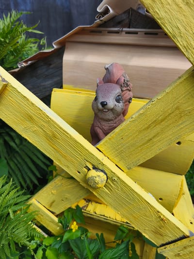This close-up outdoor shot features a small, decorative ceramic squirrel perched on a bright yellow wooden structure. The squirrel, a reddish-brown color with large eyes, is facing the camera and appears to be peeking through a gap in the yellow framing. The wooden structure is made of rough-hewn planks, painted in a vibrant yellow, with some visible wear and tear, including chipped paint and nail heads. Behind the yellow structure, we can see parts of what appears to be a wooden birdhouse or similar backyard ornament, with a light brown, slatted siding. Lush green ferns and other foliage surround the base of the structure, suggesting a garden setting. A single small yellow flower is visible amongst the greenery. The lighting suggests it could be daytime, with natural light illuminating the scene. The overall impression is one of a whimsical, garden decoration. No text is visible in the image.