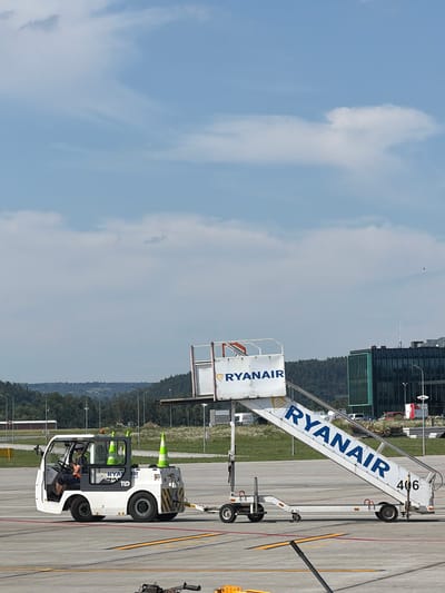 An airport tarmac scene in Balice, Poland features a white tug vehicle driven by a person wearing a yellow shirt and dark shorts, with a helmet. Two orange and white traffic cones are visible in the bed of the tug. Attached to the rear of the tug is a tow bar connected to a mobile passenger stair unit. The stair unit is white with blue lettering that reads "RYANAIR" and the number "406". In the background, a modern building with green glass windows is visible, along with trees and rolling hills under a partly cloudy sky.