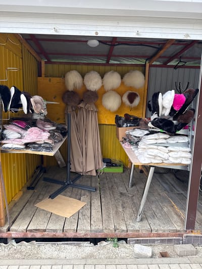 The image shows an outdoor stall with various items for sale, likely at a market. The stall is constructed with wooden planks and has a yellow corrugated metal roof and walls. In the background, a metal frame supports a tarp that is folded and leaning against the back wall.

Hanging on a rack are numerous fuzzy hats or pom-poms in various colors, including white, brown, and pink. Below these, more items are displayed, including what appear to be slippers or footwear in black and white, as well as some pink and white accessories in plastic packaging.

To the right, a metal umbrella stand is visible, with a folded tan umbrella resting on it. A piece of cardboard is propped against the wooden floor. The overall impression is that of a small retail space, possibly a temporary market stall, filled with textile goods. The scene is brightly lit, suggesting daytime, and the weather appears to be fair. There is no visible text in the image.