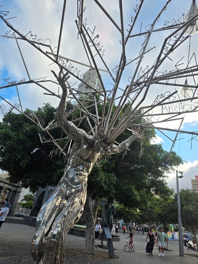 The image captures a striking outdoor sculpture in Santa Cruz de Tenerife, Spain. The sculpture, made of metallic rods and panels, depicts a human torso reaching upwards, with its arms spread wide. Branches extending from the torso are adorned with letters, forming words and phrases. In the background, lush green trees partially obscure a building with arched structures. People are seen walking and interacting in the paved public space surrounding the sculpture. A woman pushes a child on a bicycle, while other individuals stroll by. The scene is set against a backdrop of a cloudy sky. The sculpture appears to be situated in a prominent public area, likely a plaza or park, given the presence of people and urban elements.