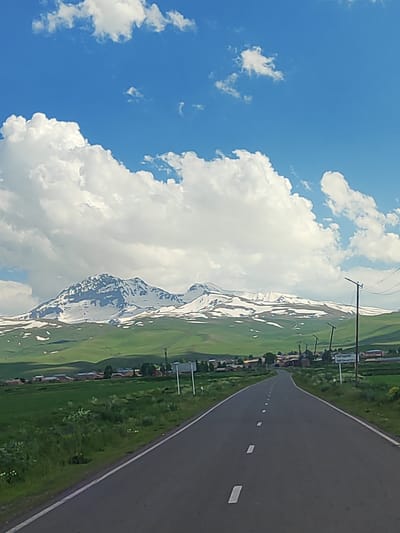The image shows a straight asphalt road leading towards a village nestled in a valley, with snow-capped mountains in the background. The sky is a vibrant blue with fluffy white clouds. To the right of the road, there is a sign with text in Armenian and Latin script, indicating "GEGHADZO". The landscape is green and hilly, with a few scattered trees and buildings visible in the village. The overall scene depicts a rural setting in Geghadzor, Armenia, under a clear, partly cloudy sky. There are no people or specific actions depicted in the image.