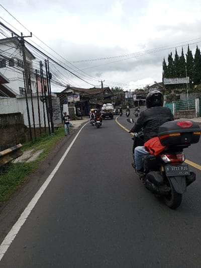 The image depicts a street scene under an overcast sky, observed from the perspective of a vehicle traveling on an asphalt road. The provided location of Taohom, Laos, is contradicted by visual evidence, specifically an Indonesian license plate ("B 3777 PJS") and Indonesian language visible on signage.
In the immediate foreground, the rear of a black Yamaha NMAX scooter is visible. The scooter is operated by a person wearing a black full-face helmet, a black jacket, and light blue jeans. A red bag is secured on the passenger seat, and a black hard case is mounted on the rear rack. The scooter's license plate reads "B 3777 PJS" with "06-26" below. The road features a solid white line on its left edge and a yellow dashed line marking the center or an adjacent lane. To the far left, a grassy verge and a rusty metal guardrail are present.
In the midground, a line of traffic proceeds ahead on the road, which curves gently to the right. This traffic includes several motorcycles and one light-colored flatbed truck carrying multiple large white sacks. On the left side of the road, a male pedestrian in a dark t-shirt and light pants walks on a dirt shoulder adjacent to the road. Behind him, a white multi-story building is partially obscured by a dense arrangement of utility poles supporting a complex network of overhead electrical and communication wires. Further along the left, signage for "CMC" and a partially visible "SUPELOT" is present on commercial buildings. On the right side of the road, a tall white sign displaying "NAGA PERDANA" in black text stands near a cluster of tall, slender evergreen trees. Another sign with a red cross symbol on a white background is visible further down the road on the right.
The background consists of additional buildings, distant tree lines, and a uniformly grey, cloudy sky.