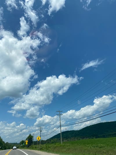 A white car drives along a rural road in Saint-André-Avellin, Canada, on a clear, sunny day with fluffy white clouds in a bright blue sky. The road is paved and has yellow and white lines indicating lanes. The car is in the left lane, heading away from the camera. To the right of the road, there are several utility poles with wires strung between them. There are also road signs, including a yellow diamond-shaped sign with a black arrow indicating a fork in the road, and a yellow circular sign with a black arrow indicating a curve to the left. Beyond the road and poles is a green field, and behind that, a tree-covered hill. The overall impression is of a peaceful drive through the countryside.