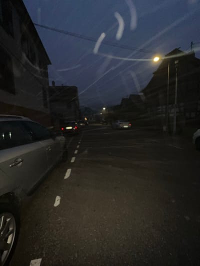 The image captures a low-light street scene in Fegersheim, France, during twilight or nighttime conditions. The asphalt road appears damp and features dashed white line markings on the left, delineating a parking area or lane, which extends towards a pedestrian crossing visible further down the road.

In the left foreground, a silver-grey vehicle, likely a car or SUV, is partially visible from its left side, with its front wheel and chrome door handle discernible. Further along the street on the left, a dark-colored vehicle displays illuminated red taillights. On the right side of the road, a lighter-colored vehicle is also visible. All visible vehicles are oriented away from the camera.

Buildings line both sides of the street. On the left, a multi-story building with multiple windows is present. On the right, structures with traditional, possibly gabled, roofs are visible, one featuring a prominent steep pitch. An illuminated streetlamp with a yellow-orange light source stands on the right side, casting light onto the scene. Faint overhead wires are visible against the dark blue sky.

No individuals are identifiable within the frame. The image exhibits prominent light streaks and flares, particularly across the upper half, and a general soft focus consistent with low-light conditions.