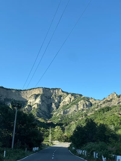 The image depicts a road winding through a mountainous, forested area under a clear blue sky. Power lines are visible stretched across the sky. The road is paved and has white dashed lines marking the lanes. On the left side of the road, there's a utility pole and dense green foliage. The background is dominated by large, rocky mountains with patches of green vegetation. A small red-roofed building can be seen nestled among the trees on the mountainside. The scene appears to be a rural or natural landscape, possibly on the outskirts of a town or village in Georgia, given the mention of Georgia in the prompt. No people or animals are visible in the image.