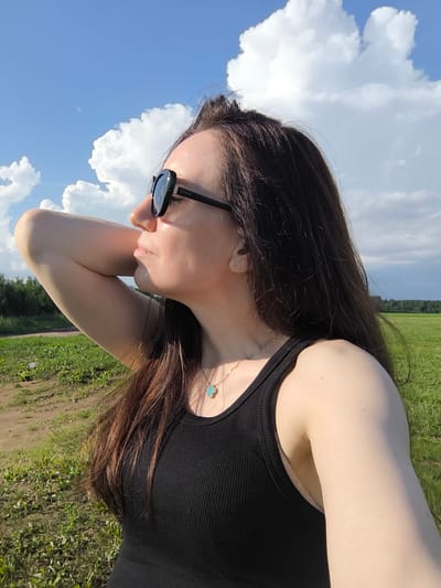 A medium shot captures a woman in profile, looking to the left, with her right arm bent and resting behind her head. She wears sunglasses and a black ribbed tank top, with a delicate gold chain necklace featuring a turquoise clover pendant. Her long, dark brown hair is loose and falls over her shoulder. The background is a grassy field under a partly cloudy blue sky, suggesting an outdoor setting. The overall lighting indicates it is daytime, likely late afternoon due to the angle of the sun casting a highlight on her face and arm. There is no indication of the specific location within the city of Vētras, Latvia.