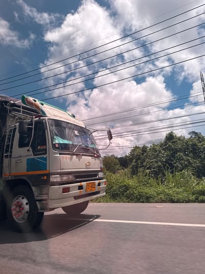 The image shows the front left side of a light-colored truck traveling on a paved road. The truck has a license plate that reads "72-4332" and "THAILAND 92". There is a ladder on the driver's side of the truck, and the word "RANGER" is visible on the side of the cab. Above the truck, power lines are strung across the frame, with a cloudy blue sky in the background. Lush green foliage and trees line the right side of the road. The scene appears to be captured from a moving vehicle, indicated by the motion blur of the road and the angle of the truck. The location is identified as Ban Krasom, Thailand.