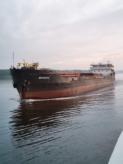 A large, dark-colored cargo ship, the "VOLGO-BALT 136," sails on a calm body of water, likely a river or canal, under an overcast sky. The ship's bow is pointed towards the left side of the frame, showing its anchor and a white wake trailing behind as it moves. The ship has a long, flat deck with visible cargo or loading mechanisms. A multi-story white superstructure is located at the stern of the ship. The water reflects the ship and the sky, creating a slightly distorted image. The surrounding environment includes a tree line visible on the distant left bank of the waterway. The image appears to have been taken from another vessel or the shore. The location is identified as Ratibor, Russia.