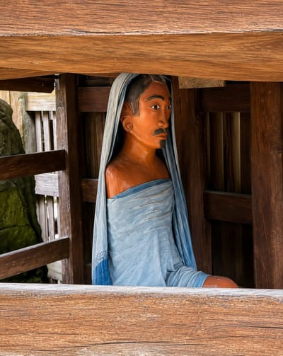 A carved wooden statue of a man, with orange skin and a blue cloth draped around him, is seated within a wooden structure. The man has a stern expression, with dark hair styled with a comb-like pattern, and a thin mustache and goatee. The statue is positioned under a wooden beam and in front of vertical wooden planks. To the left of the statue, a large, moss-covered rock is partially visible, along with a wooden fence. The overall scene appears to be an outdoor setting within the historical village of Ambarita, Indonesia, likely part of the Ambarita Stone Chairs site, which is known for its ancient Batak stone furniture and artifacts.