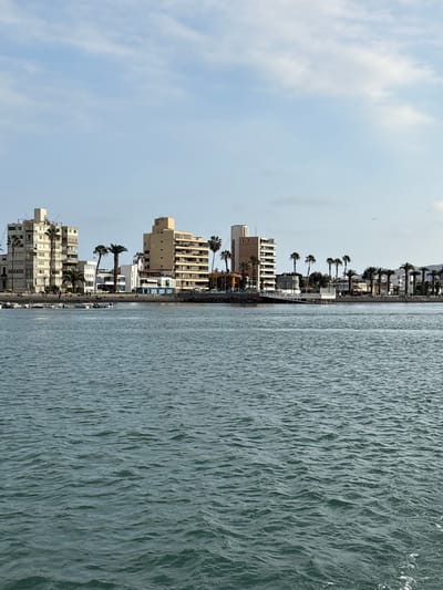 The image shows a waterfront view of the city of La Punta, Peru. In the foreground, there is a body of water with small waves. In the background, there is a shoreline with buildings, palm trees, and a beach. Several small boats are moored near the shore. People can be seen walking on the beach and near the buildings. The sky is partly cloudy.