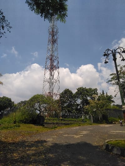A daytime image captured in La Roche-en-Ardenne, Belgium, depicts a tall, self-supporting lattice telecommunications tower positioned left-of-center, extending from the mid-ground into a partly cloudy blue sky. The tower is silver-grey with distinct red-painted sections. Its upper portion is partially obscured by dense green tree foliage originating from the left foreground.
The foreground and mid-ground consist of an irregularly shaped, dark asphalt or paved surface, strewn with numerous dry, dark leaves. Patches of green grass are visible along the edges of the pavement. Additional green trees and bushes surround the base of the tower and line the mid-ground. A low stone or concrete retaining wall is visible behind vegetation on the right mid-ground. A decorative streetlamp post, dark in color with multiple ornate arms, is partially visible on the far right, extending upward into the frame.
A single individual, a child, is observed on a bicycle on the paved surface at the far right edge of the frame, moving away from the viewer. No other people or direct interactions are visible. The scene is illuminated by strong natural sunlight, casting discernible shadows on the ground.