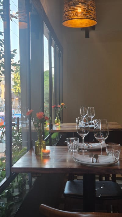 The image depicts the interior of a restaurant in Rehovot, Israel, viewed through a large window that offers a glimpse of the city's streetscape. Inside, two wooden tables are set for dining. The table in the foreground is adorned with two green glass bottles, a small lit candle in a glass holder, and two glasses. The table in the background is more elaborately set with multiple wine glasses, plates, napkins, and cutlery. Several small vases with orange and yellow flowers are placed on both tables, adding a touch of color. A warm, ambient light emanates from a woven lampshade hanging from the ceiling. Outside the window, trees and buildings are visible, along with a blurred street scene that suggests moderate urban activity. The overall atmosphere is inviting and suggests a dining experience in a restaurant setting.
