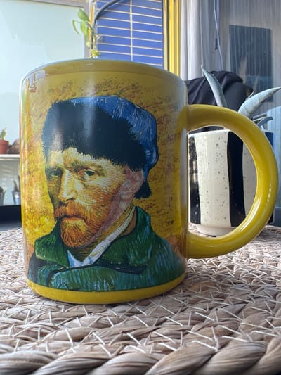 The image shows a yellow ceramic mug with a depiction of Vincent van Gogh's self-portrait. The mug is placed on a woven placemat, likely made of straw or a similar material. In the background, partially visible, is another mug with black and white vertical stripes. Behind the mugs, there are vertical blinds and some foliage from plants. The lighting suggests an indoor setting, possibly a kitchen or dining area, with natural light coming from a window. The scene does not provide any specific indicators to pinpoint its location within the city of Mollet del Vallès, Spain.