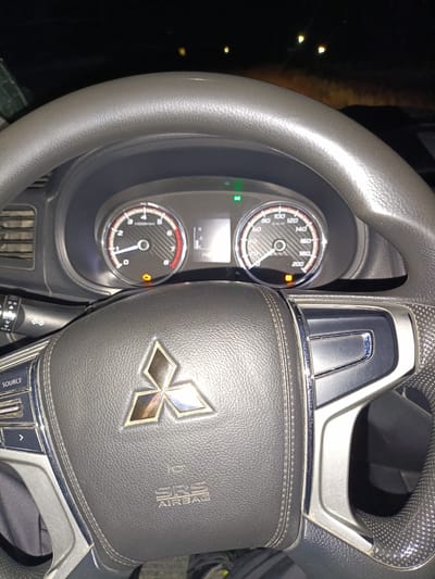 The image shows the interior of a vehicle, specifically the steering wheel and dashboard. The steering wheel is black leather with silver accents and features the Mitsubishi logo in the center. On the dashboard, there are two circular gauges, one appearing to be a tachometer and the other a speedometer. An orange indicator light is illuminated on the speedometer. A turn signal lever is visible to the left of the steering wheel. The scene is dimly lit, suggesting it is nighttime. There is no indication of a specific location within a city or country.