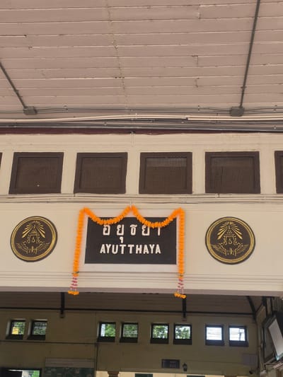 The image shows a building in Phra Nakhon Si Ayutthaya City Municipality, Thailand, with a white facade and dark brown trim. The word "AYUTTHAYA" is written in English and Thai characters on a dark background in the center of the building. There are two gold circular emblems above and below the sign. An orange garland hangs around the sign. To the right of the facade, there's a glimpse into the interior of the building with a view of windows and an "EXIT" sign. The facade has rows of small, square, dark-colored vents. The leftmost part of the image shows a wall with electrical conduits and a junction box.