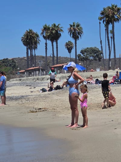 The image depicts a sunny day at San Clemente State Beach in San Clemente, California, USA. In the foreground, a woman in a light blue bikini and a white cap stands with her hands on her hips, looking to the left. Beside her, a young girl in a pink swimsuit holds a piece of sand. To their right, a young boy in a black shirt and patterned swim trunks stands facing away. In the background, several people are scattered across the sandy beach, some lying on towels or sitting in chairs, under palm trees and a blue umbrella. The shoreline features pebbles and dry seaweed. Further back, a row of buildings with orange roofs and solar panels line the edge of the beach, with a hilly, vegetated landscape beyond.