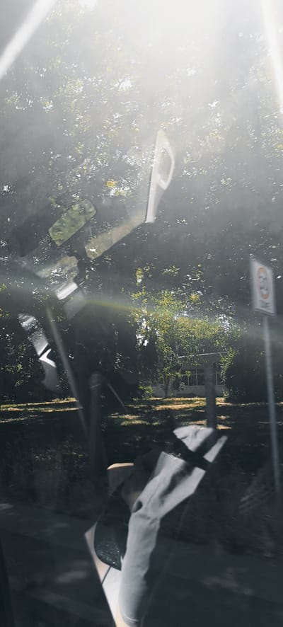 The image appears to be taken from inside a vehicle, looking out through the windshield or side window. The view is dominated by bright sunlight filtering through a dense canopy of green trees, creating lens flares and a hazy, overexposed effect in the upper portion of the frame.
In the foreground, several reflective surfaces, likely parts of the vehicle's interior such as the rearview mirror, are visible and create distorted reflections of the outside environment. There are also some abstract, light-colored shapes that may be parts of the car's dashboard or other interior components.
Further out, through the greenery, a small, light-colored building with several windows is visible. To the right of the building, a traffic sign is partially visible. It is a circular sign with a red border and a white background, featuring a black silhouette of a vehicle. Inside the circle, there is a red bar across the top. Below the circle, there appears to be text, possibly indicating a speed limit.
The ground visible beneath the trees appears to be a mix of dry grass and dirt. The overall impression is a sunny day, possibly in a semi-rural or suburban area with vegetation, and the presence of the traffic sign suggests a road or public area. The lens flares and reflections indicate that the sun is positioned behind the viewer, or at an angle that causes significant glare.
It is not possible to definitively identify the location as Winterbach, Germany, based solely on the visual information in this image.