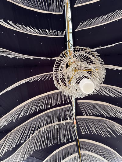 A low-angle shot showcases a black fabric ceiling adorned with curvilinear designs of white fabric strips ending in frayed strands. A round, modern chandelier hangs centrally, constructed with tiered, translucent elements and a white globe at its core. A structural beam is visible through an opening in the fabric. The ceiling fabric is also dotted with small, sparse points.