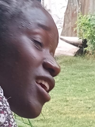The image shows a close-up, profile view of a young Black person's face. Their eyes are partially closed, and their mouth is open, revealing their teeth. The person is wearing a floral-patterned garment on their left shoulder. The background consists of green grass and some indistinct structures, including what appears to be a white, irregular shape on the left and a textured, vertical surface on the right. A bush with green leaves is visible in the upper right quadrant. The lighting suggests an outdoor setting during daylight. There are no discernable landmarks that would indicate a specific location within Mbar, Nigeria.