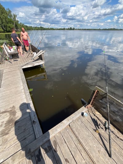 This media file depicts a scene of leisure on a wooden dock by a calm body of water under a bright, cloudy sky. Two individuals are present on the dock, engaged in fishing. One man, shirtless and wearing red shorts, is actively fishing. The second person, seen from the back, is wearing dark shorts and a t-shirt, and is also near fishing rods. Several fishing rods are set up on the dock, with lines cast into the water.
The setting appears to be a natural, rural environment, given the presence of trees lining the far bank of the water. The time of day is clearly daytime, suggested by the strong sunlight casting shadows on the wooden planks. The weather is fair, with a mix of sun and clouds visible in the sky, which are reflected in the water's surface.
Notable objects on the dock include fishing equipment, a small metal bucket, and personal items near the individuals. The overall mood conveyed is one of relaxation and enjoyment of outdoor activities. The presence of the dock and the surrounding greenery suggests a location such as a lake or a wide river. The context provided indicates this scene is located in Вяззеўскі сельскі Савет, Belarus. There is no visible text in the image.