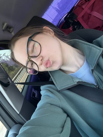 A young woman is seated in the driver's seat of a car, presumably traveling along a road near Pesochny, Russia. She is angled sideways, facing the camera with her head tilted. She wears glasses and a light-blue top, zipped into a green hoodie. A dark-colored seatbelt crosses her body. A red bag is positioned behind her. The background shows blurred trees and road markings visible through the car window, suggesting movement.