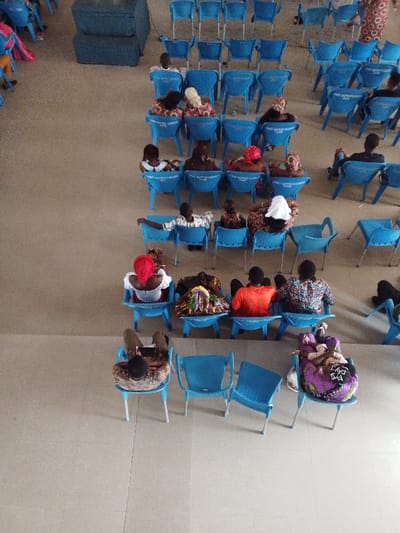 The image shows a crowded interior space with rows of blue plastic chairs arranged in a rectangular configuration. The scene is likely a church or a community hall in Yenagoa, Nigeria. The image captures a moment during a gathering or event, with people seated and engaged in conversation. Some individuals are engaged in conversation while others are observing or listening attentively. The individuals are diverse in age and attire, reflecting the local cultural context. A couple of individuals are seated on a blue chair in the foreground. The people in the background are blurred, showcasing their movement and interaction. The image is taken from a high angle, offering a panoramic perspective of the scene. The chairs are neatly arranged, indicating a formal setting. The environment appears clean and well-maintained, suggesting a sense of order and organization. The image reveals a glimpse into a social gathering in Yenagoa, showcasing the community's dynamic and engaging nature.