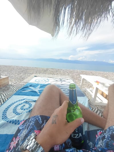 A first-person perspective shot captures a person relaxing on a beach blanket under a thatched umbrella. The person's hand holds a green bottle of "EZA LAGER" beer. The individual is wearing floral swim trunks and a watch. In the background, a pebble beach meets a calm, light blue sea. Distant mountains form the horizon under a partly cloudy sky. A white beach chair and a small wooden crate are also visible on the beach. The scene suggests a leisurely day at the shore, possibly in the Municipal Unit of Diakopto, Greece, known for its coastal areas and proximity to mountainous landscapes.