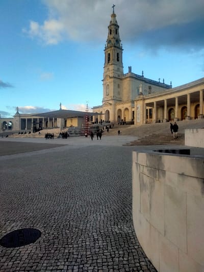 This image displays the Sanctuary of Fátima in Fátima, Portugal, under a partly cloudy, blue sky. The scene is illuminated by natural light, casting long shadows indicative of late afternoon conditions.
In the foreground, the ground is covered with dark gray cobblestone pavement, featuring a circular black manhole cover in the lower left. A curved, light-colored concrete or stone wall partially frames the lower right portion of the image.
The mid-ground and background reveal the architectural complex of the Sanctuary. Dominating the right side is the Basilica of Our Lady of Fátima, a large structure constructed from light-toned stone, featuring a tall, ornate bell tower topped with a cross and equipped with a clock face. To the left of the Basilica, the modern Basilica of the Most Holy Trinity is visible, characterized by its wide, flat-roofed canopy supported by columns, under which an altar is present.
The expansive plaza, known as the Esplanade of the Sanctuary, is bordered by multi-tiered steps and colonnades with arches, also built from light-colored stone. Several statues are positioned atop sections of the buildings and colonnades.
Approximately 20-30 individuals are scattered across the plaza and on the steps, engaged in walking or standing. They are dressed in dark, heavy clothing such as coats and jackets. A distinctive red, conical, multi-level structure, resembling a contemporary Christmas tree, stands in the central plaza area.