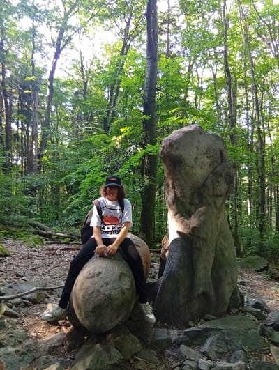 A person with medium-length curly hair, wearing a white t-shirt with an orange graphic, black pants, a black baseball cap, and sneakers, is sitting on a large round boulder in a forest. The person is looking towards the camera. To the right of the person is a tall, irregularly shaped rock formation. The ground is covered with rocks, dirt, and fallen leaves. Tall trees with green foliage form a canopy overhead, allowing sunlight to filter through. The scene appears to be the "Małpka" (Little Monkey) rock formation located in the Góra Ślęża National Park near the town of Sobótka, Poland.