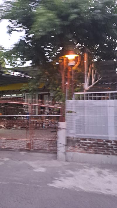 The image depicts a street scene in Lhokawe Teungoh, Indonesia. A streetlamp with an orange glow is visible amidst lush green foliage. In the background, a building with a yellow roof and a brick wall is partially obscured by a white, slatted fence. A rusty metal fence runs along the foreground, and the ground appears to be paved or concrete. The overall impression is a snapshot of a residential or commercial area, likely taken during dusk or evening given the illuminated streetlamp.